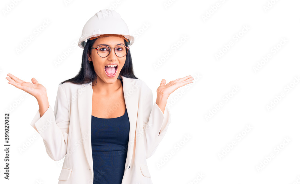 Young beautiful latin girl wearing architect hardhat and glasses celebrating crazy and amazed for success with arms raised and open eyes screaming excited. winner concept Young beautiful latin girl wearing architect hardhat and glasses celebrating crazy and amazed for success with arms raised and open eyes screaming excited. winner concept