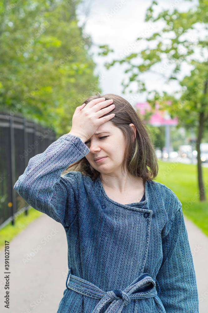 A girl holds her head while standing on the street against the backdrop of a park fenced in. Concept - headache and general fatigue