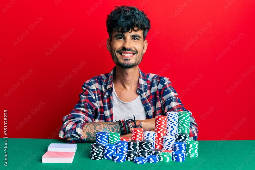 Young hispanic man sitting on the table with poker chips and cards happy face smiling with crossed arms looking at the camera. positive person. Young hispanic man sitting on the table with poker chips and cards happy face smiling with crossed arms looking at the camera. positive person.