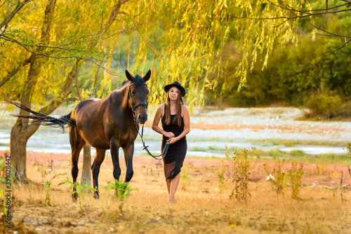 Fototapeta Naklejka Na Ścianę i Meble -  A girl in a beautiful black dress and a black hat walks with a horse across the field