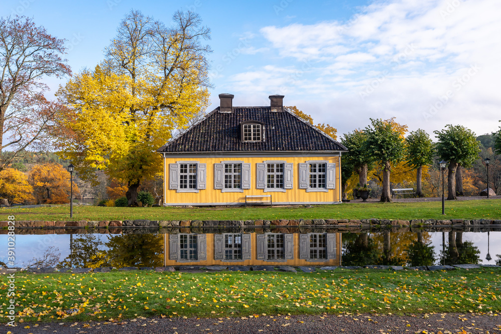 Small cozy old house of the past centuries with 4 windows and a tiled roof. A yellow house in the countryside is reflected in the water on a sunny autumn day. Country villa cottage in vintage style.