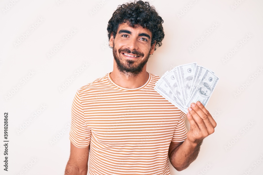 Handsome young man with curly hair and bear holding money bag with dollar symbol looking positive and happy standing and smiling with a confident smile showing teeth Handsome young man with curly hair and bear holding money bag with dollar symbol looking positive and happy standing and smiling with a confident smile showing teeth
