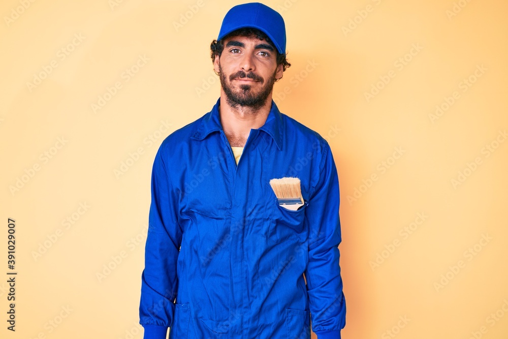 Handsome young man with curly hair and bear wearing builder jumpsuit uniform relaxed with serious expression on face. simple and natural looking at the camera.