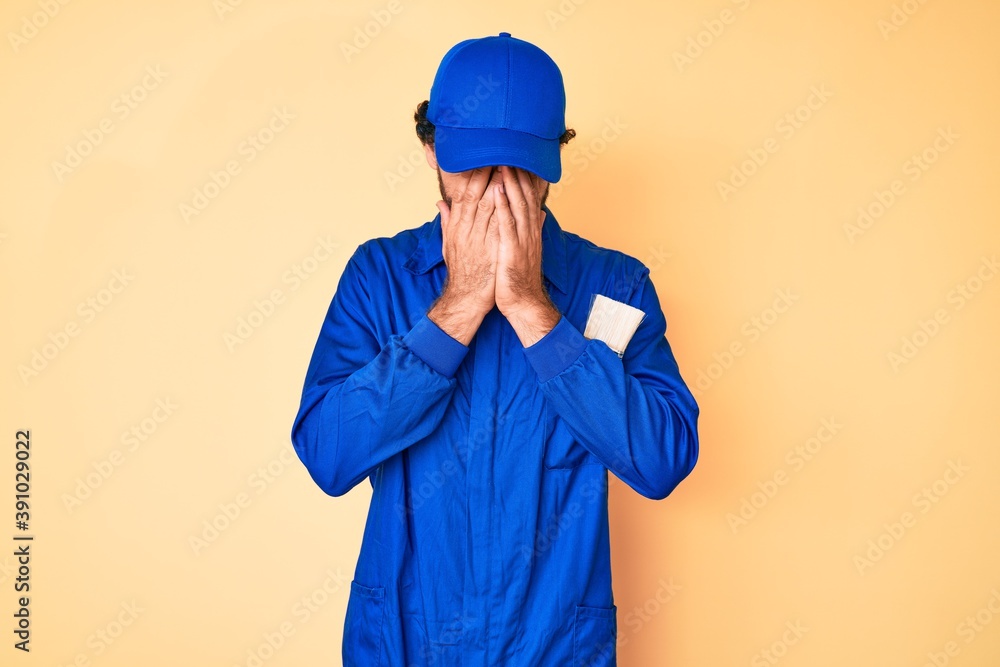 Handsome young man with curly hair and bear wearing builder jumpsuit uniform with sad expression covering face with hands while crying. depression concept.