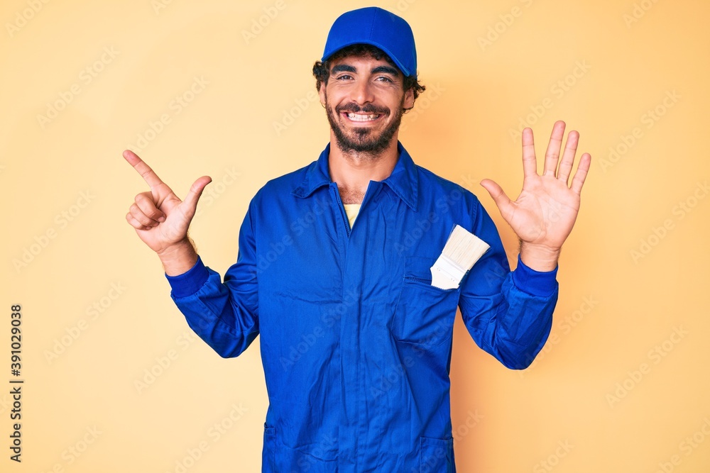 Handsome young man with curly hair and bear wearing builder jumpsuit uniform showing and pointing up with fingers number seven while smiling confident and happy.