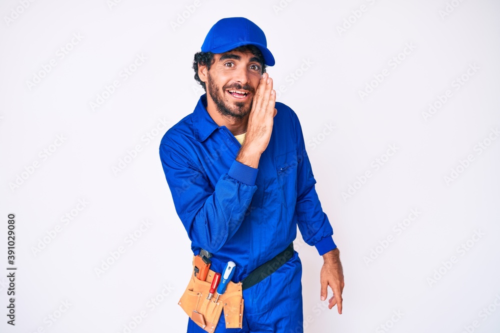 Handsome young man with curly hair and bear weaing handyman uniform hand on mouth telling secret rumor, whispering malicious talk conversation Handsome young man with curly hair and bear weaing handyman uniform hand on mouth telling secret rumor, whispering malicious talk conversation