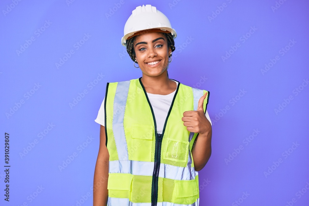 Young african american woman with braids wearing safety helmet and reflective jacket smiling happy and positive, thumb up doing excellent and approval sign