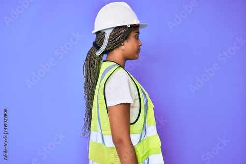 Young african american woman with braids wearing safety helmet and reflective jacket looking to side, relax profile pose with natural face with confident smile.