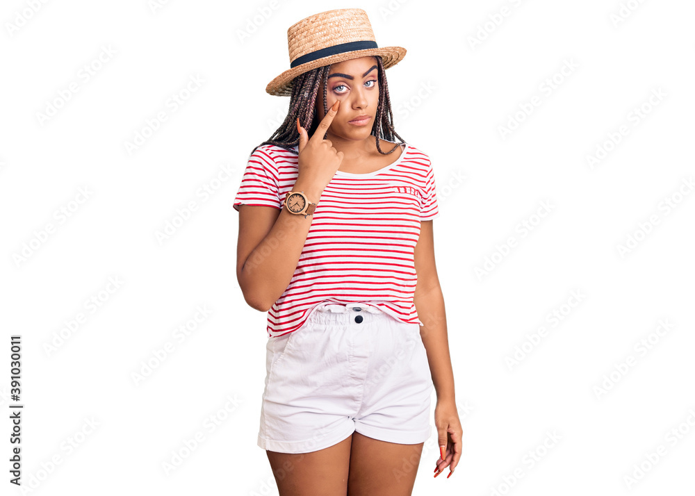 Young african american woman with braids wearing summer hat pointing to the eye watching you gesture, suspicious expression