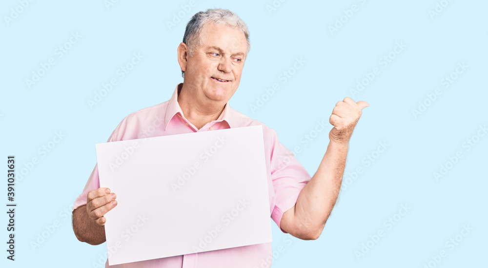 Senior grey-haired man holding blank empty banner pointing thumb up to the side smiling happy with open mouth Senior grey-haired man holding blank empty banner pointing thumb up to the side smiling happy with open mouth