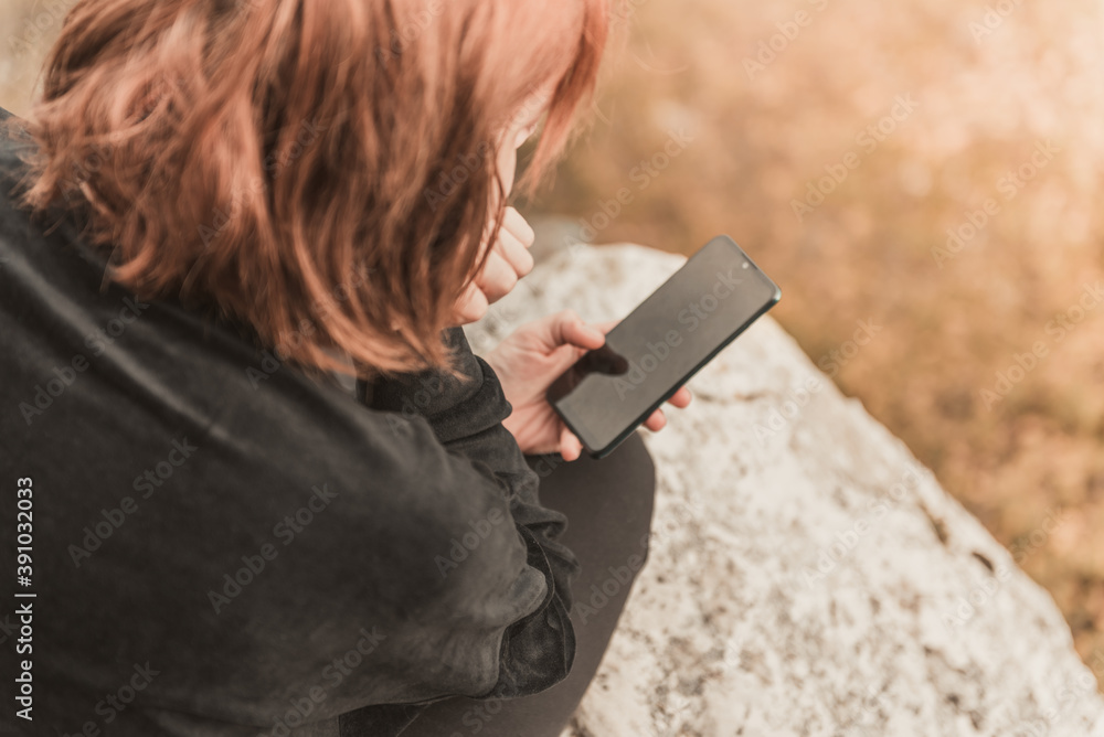 The red-haired traveler girl writes SMS on her smartphone sitting on the mountain rock. The concept of travel, technology. The red-haired traveler girl writes SMS on her smartphone sitting on the mountain rock. The concept of travel, technology.