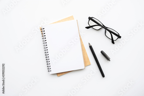 Blank notebook is on top of white office desk table with pen and eye glasses. Top view, flat lay.