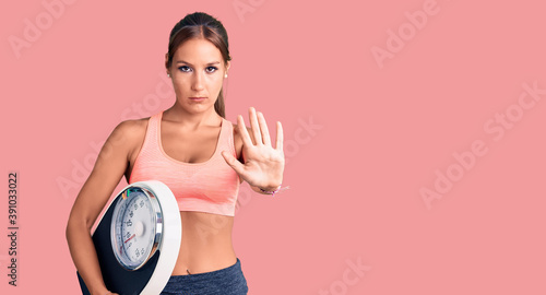 Young beautiful hispanic woman wearing sportswear holding weighing machine with open hand doing stop sign with serious and confident expression  defense gesture