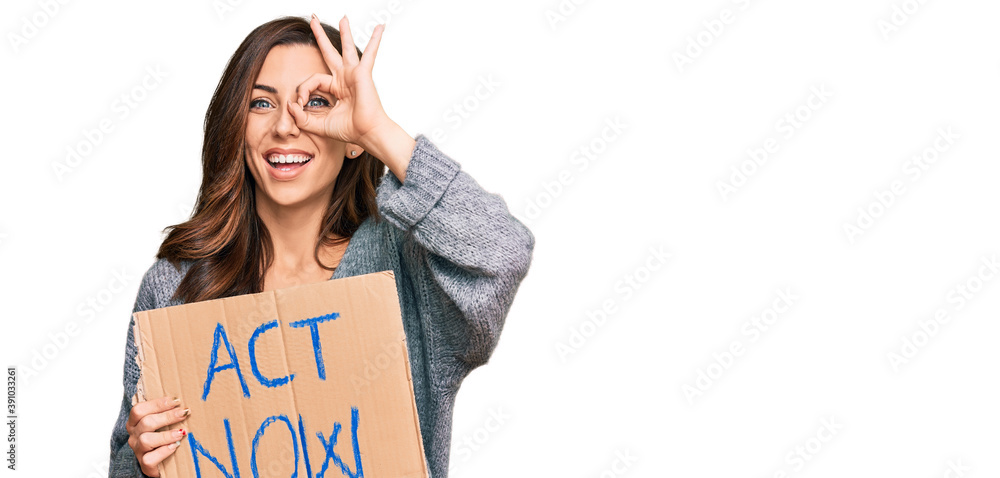 Young brunette woman holding act now banner smiling happy doing ok sign with hand on eye looking through fingers Young brunette woman holding act now banner smiling happy doing ok sign with hand on eye looking through fingers