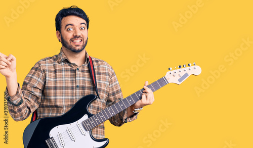 Young hispanic man playing electric guitar smiling happy pointing with hand and finger to the side