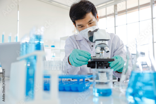 Young Asia male scientist looking through the lens of a microscope while being seated inside of a laboratory.