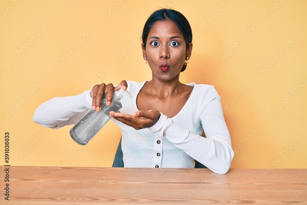 Young indian girl sitting on the table cleaning hand using sanitizer gel making fish face with mouth and squinting eyes, crazy and comical. Young indian girl sitting on the table cleaning hand using sanitizer gel making fish face with mouth and squinting eyes, crazy and comical.