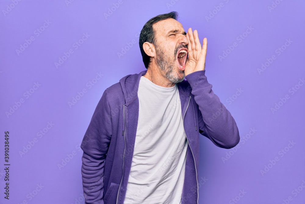 Middle age handsome sporty man wearing casual purple sweatshirt over isolated background shouting and screaming loud to side with hand on mouth. Communication concept. Middle age handsome sporty man wearing casual purple sweatshirt over isolated background shouting and screaming loud to side with hand on mouth. Communication concept.