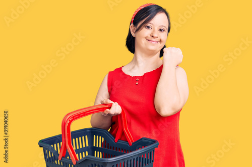 Brunette woman with down syndrome holding supermarket shopping basket screaming proud, celebrating victory and success very excited with raised arms photo