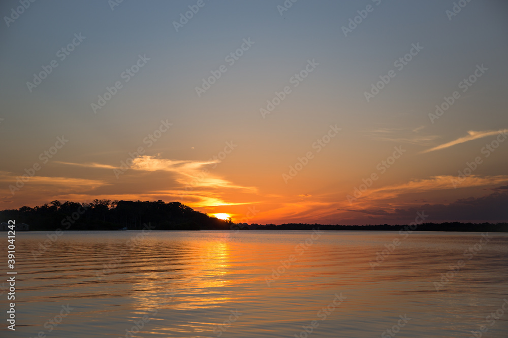 Beautiful Amazon River sunset over horizon with the sun setting behind jungle trees & forest foliage & reflecting into the Amazonian water in the State of Amazonas, Brazil, South America