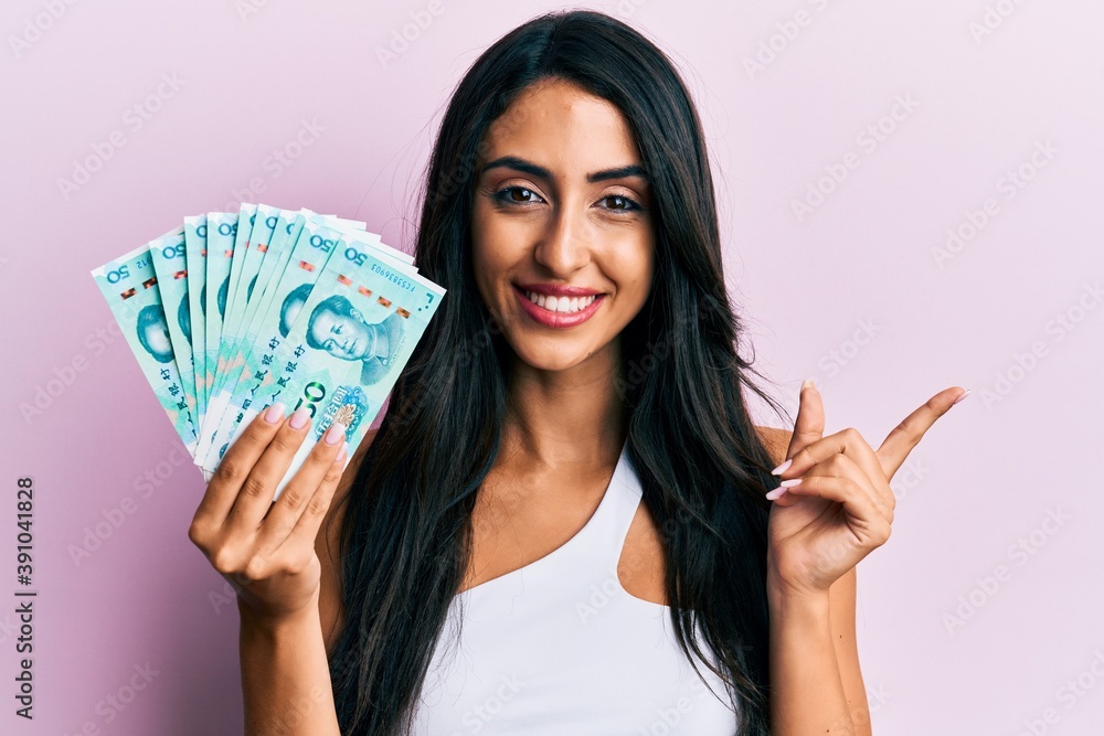 Beautiful hispanic woman holding yuan chinese banknotes smiling happy pointing with hand and finger to the side Beautiful hispanic woman holding yuan chinese banknotes smiling happy pointing with hand and finger to the side