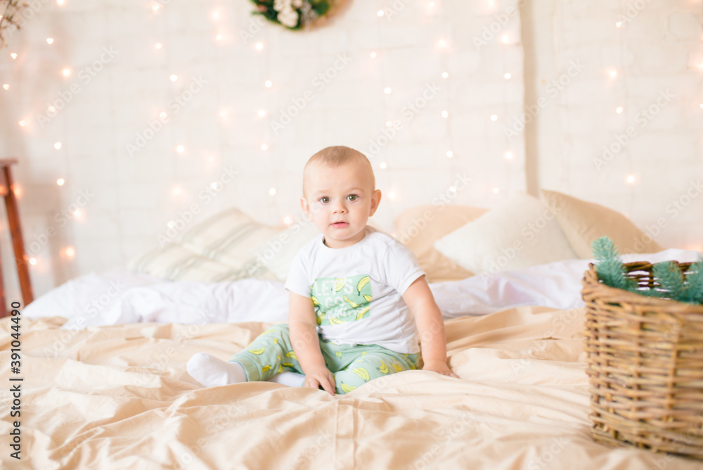 Fototapeta premium Little boy in pajama have fun in in a Scandinavian style bedroom decorated with Christmas garlands and needles on a large bright bed. Christmas mood. Christmas mood. Family time 