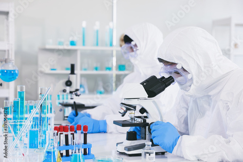 A medical professional in an antiplague suit is fully protected from the virus and wear sterile rubber gloves and looking through the lens of a microscope while being seated inside of a laboratory.