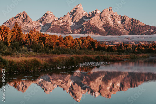 Sunrise glow hits the peaks and trees of the Teton Range of the Rocky Mountains. Schwabacher Landing in Grand Teton National Park, WY, USA. Water reflections of the Teton Range on the Snake River. 