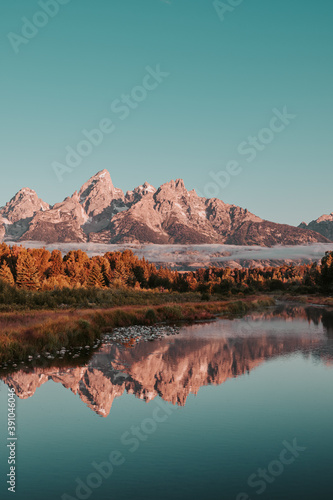 Sunrise glow hits the peaks and trees of the Teton Range of the Rocky Mountains. Schwabacher Landing in Grand Teton National Park, WY, USA. Water reflections of the Teton Range on the Snake River. 