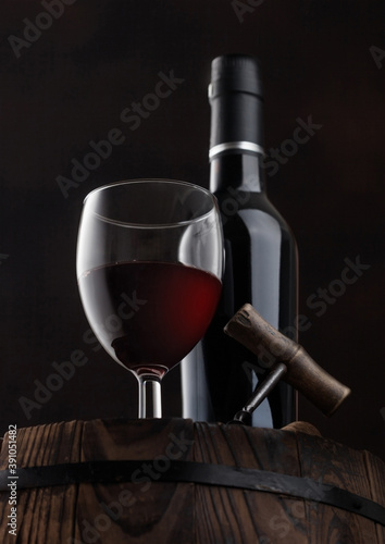 Bottle and glass of red wine with corks and vintage corkscrew on top of wooden barrel on black.