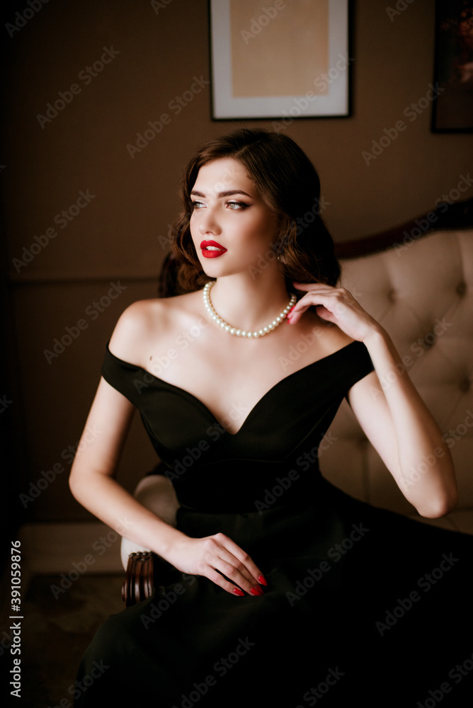 Lovely young woman in black evening dress and pearl necklace, classic  make-up in the style of Coco Chanel, on a sofa in a classic interior.  Beauty and fashion. Stock-foto