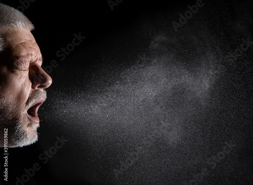 A man sprays aerosols into the air while speaking. The background is black.