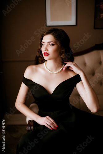 Lovely young woman in black evening dress and pearl necklace, classic make-up in the style of Coco Chanel, on a sofa in a classic interior. Beauty and fashion. photo