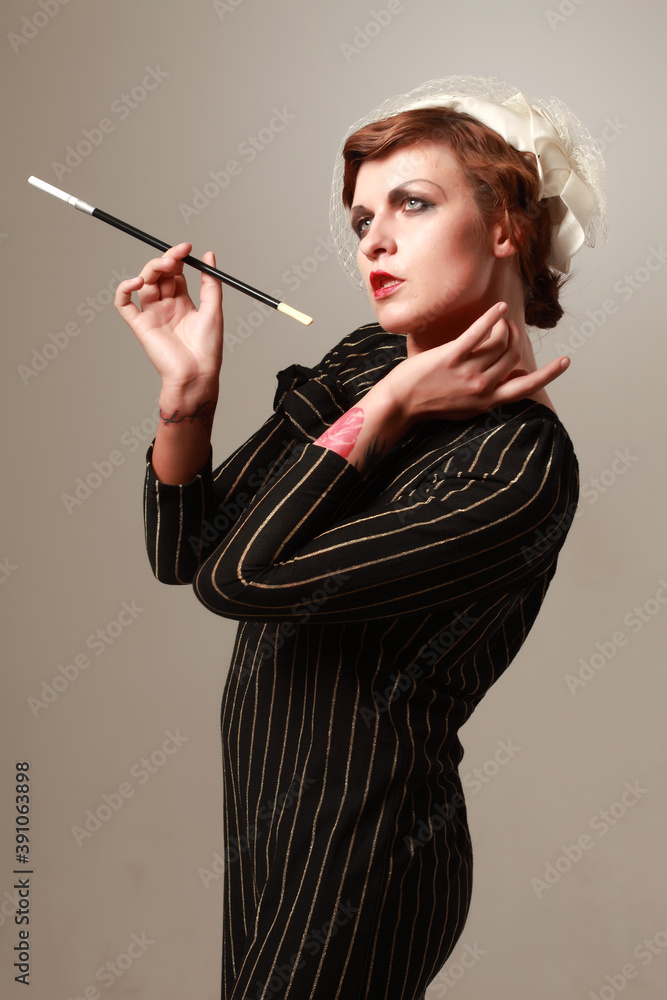 Obraz premium Studio portrait of young female model with vintage makeup and dress posing with an old fashioned cigarette against grey background