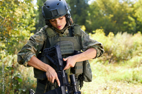 confident soldier woman loading rifle in the forest, alone, looking at gun, wearing military wear