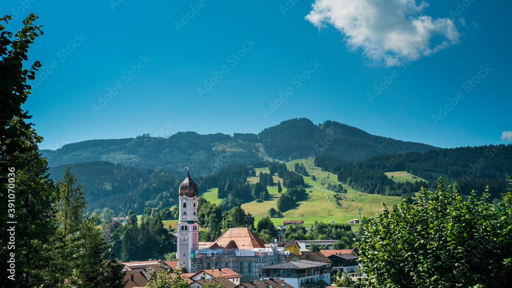 Germany, Allgaeu, Nesselwang city church building and houses with alpspitz mountain and cable car transportation under blue sky