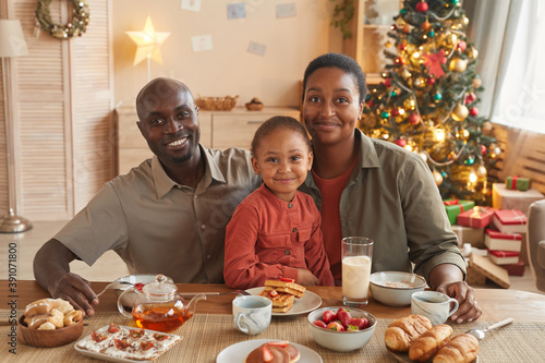 Portrait of happy African-American family enjoying tea and sweets while celebrating Christmas at home in cozy home interior