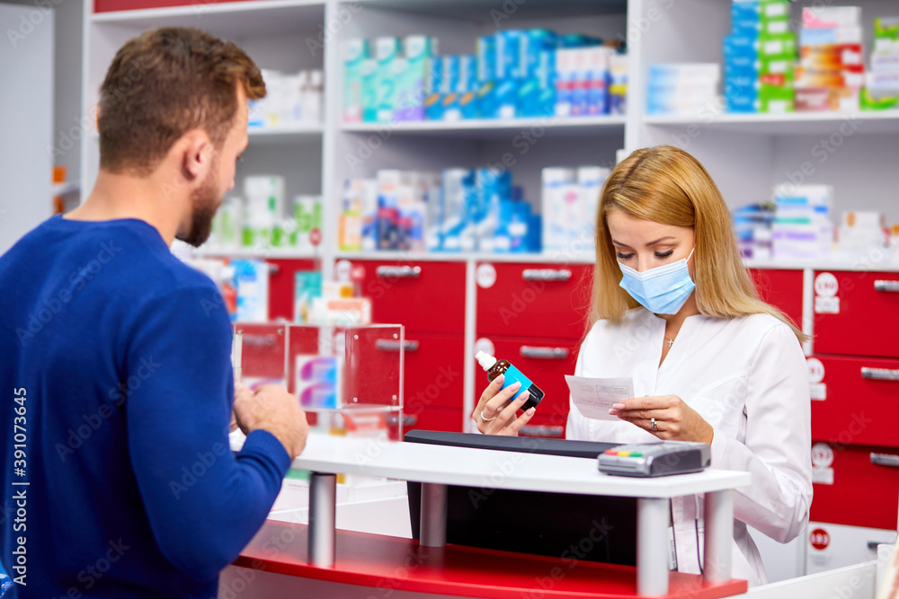 young beautiful caucasian pharmacist in protective medical mask is working with customer in modern drugstore, serve the client, wearing white medical gown