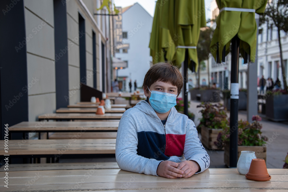 a boy wearing a mask because of the coronavirus sits in a street cafe a boy wearing a mask because of the coronavirus sits in a street cafe