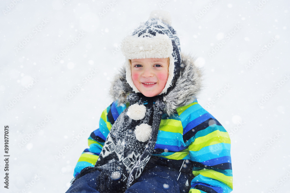 a small child boy in a bright striped jacket sitting on a sledge smiles against the background of snowfall. Winter entertainment a small child boy in a bright striped jacket sitting on a sledge smiles against the background of snowfall. Winter entertainment
