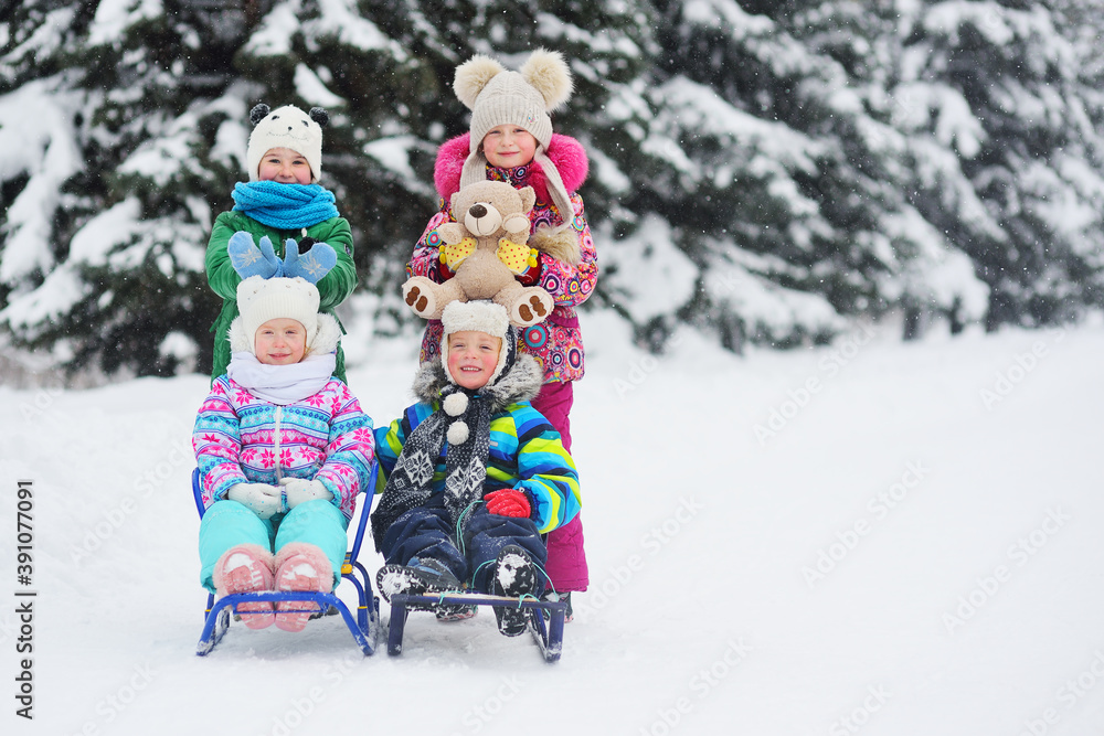 a group of children in bright colored winter clothes on a sled with toys in their hands smile and play against the background of snow, snowfall and winter forest