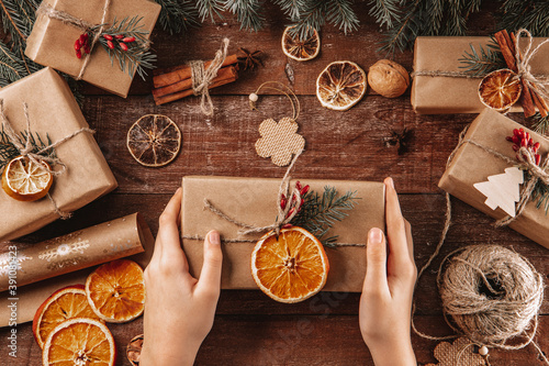  Hands hold a Christmas gift wrapped in kraft paper and natural materials. Green new year concept