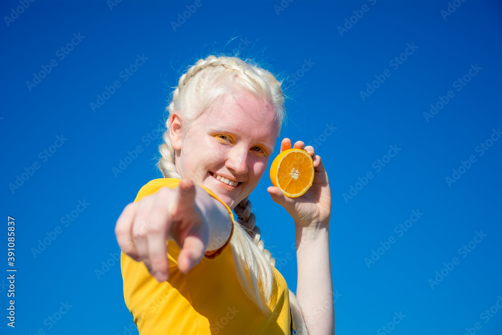 A young albino woman points a finger at the camera and holds a slice of orange in her other hand. against the blue sky. A young albino woman points a finger at the camera and holds a slice of orange in her other hand. against the blue sky.