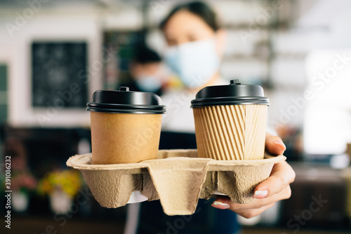 Woman Barista wearing a face mask serving coffee in takeaway paper disposable cups in the coffee shop. Small green business during coronavirus pandemic. Owner and employee service for the client.
