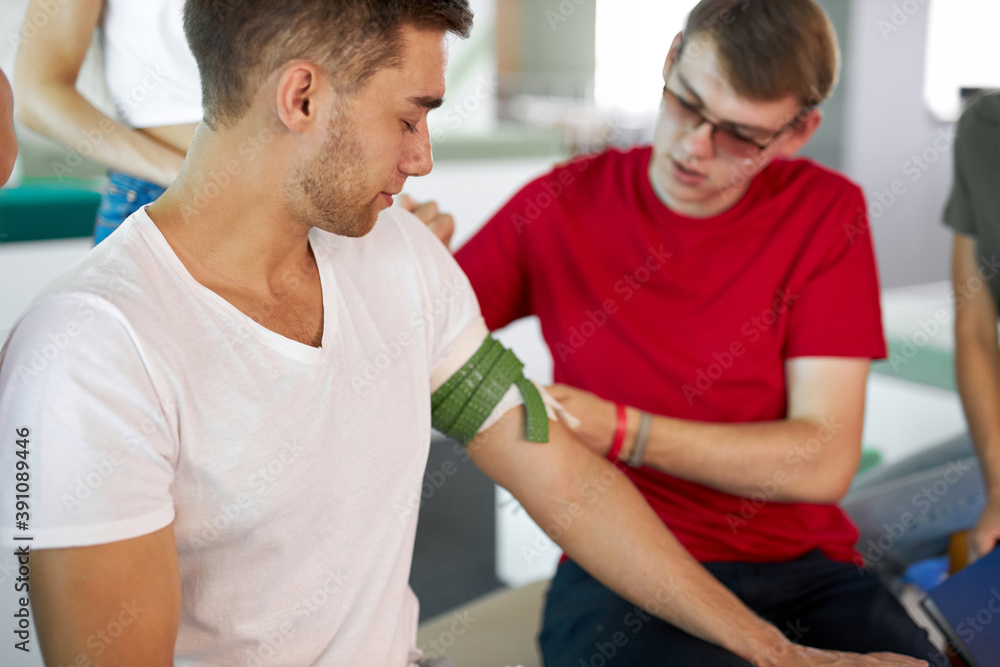 Obraz premium man wearing tourniquet on arm of injured person during class of giving first aid, help people. indoors