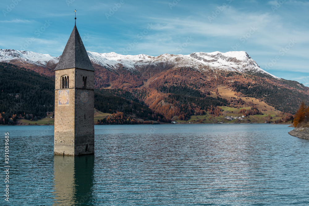Famous submerged church steeple arising from the alpine Resia Lake, in the town of Curon Venosta, in the italian Dolomite region of Trentino Alto Adige. Water rippling, sunny day. Scenic view.