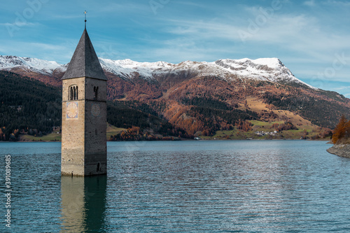 Famous submerged church steeple arising from the alpine Resia Lake, in the town of Curon Venosta, in the italian Dolomite region of Trentino Alto Adige. Water rippling, sunny day. Scenic view.