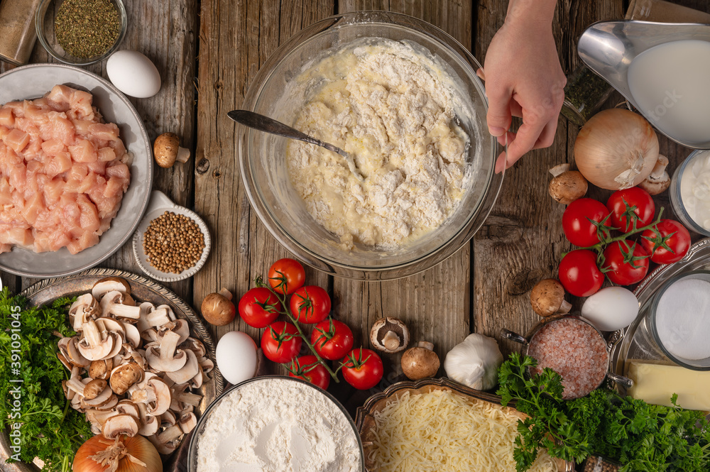 Fototapeta premium Glass bowl with dough for pie on wooden rustic table with variety of ingredients background. Concept of cooking process. Backstage of preparing tasty meal. View from above. Flat lay.