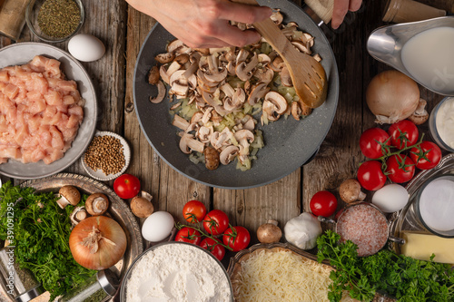 Chef stir frying mushrooms in the pan on wooden table with variety of ingredients background. Concept of cooking process. Backstage of preparing tasty meal. View from above. Flat lay.