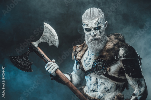 Armed with two handed axe northern dead warrior with naked torso and pale skin poses in dark smokey background.